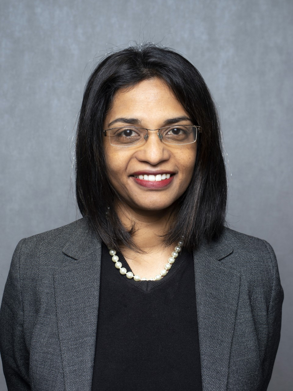 A portrait photograph of Professor Kathreena Kurian. Dark haired woman wearing glasses and smiling, grey background